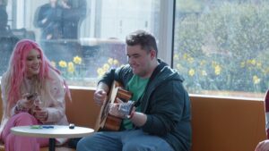 Young woman with pink hair sitting beside young man playing the acoustic guitar.