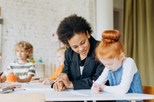 Adult woman sitting beside and assisting a young girl in a classroom setting.